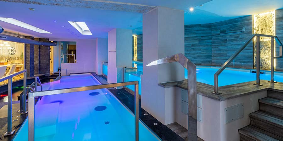 Image of hotel spa area with two small pools one on a lower level to the left and another pool on a higher level on the right. A window into the higher pool can be seen looking into the water from the lower pool. With relaxing coloured hued lighting. 